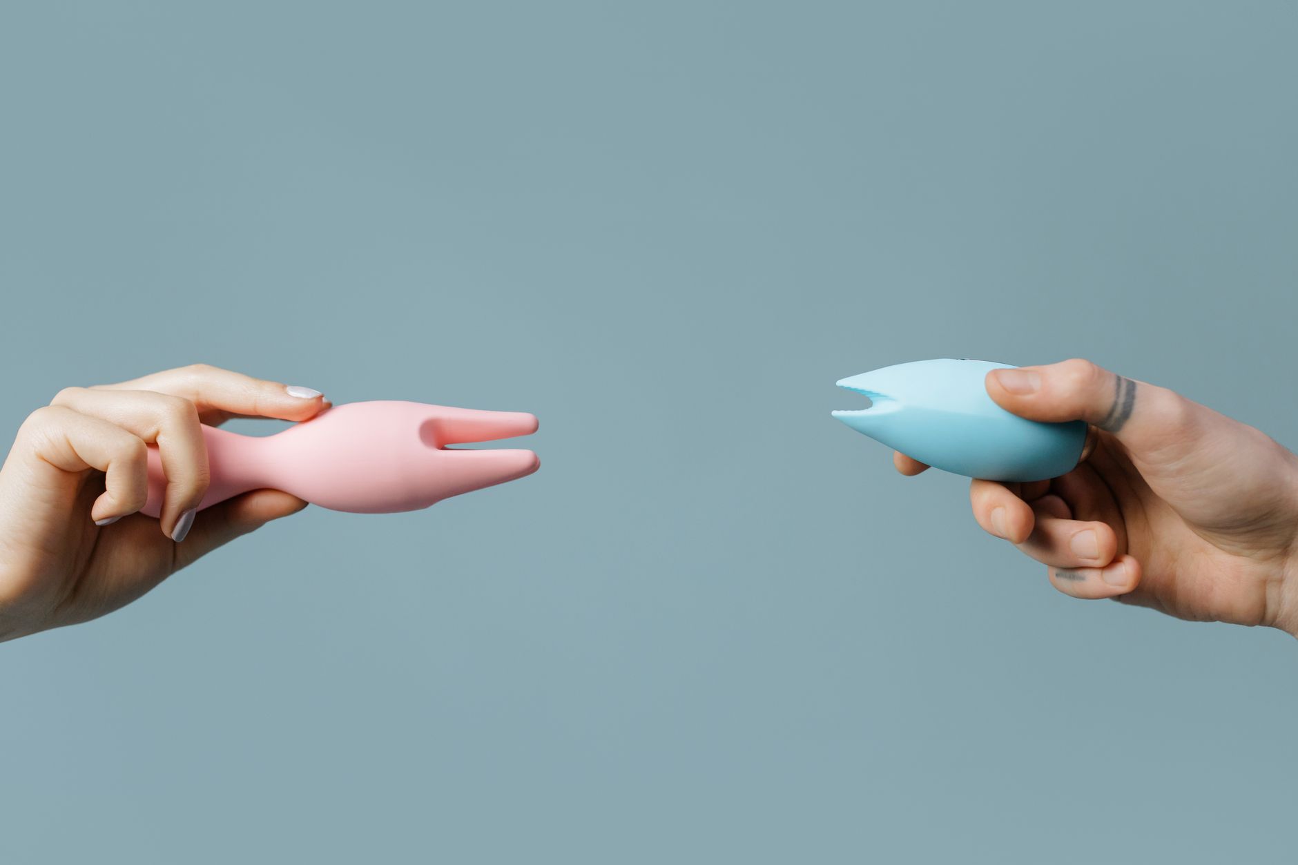 Deux mains tenant des vibromasseurs en silicone rose et bleu contre un fond pastel.