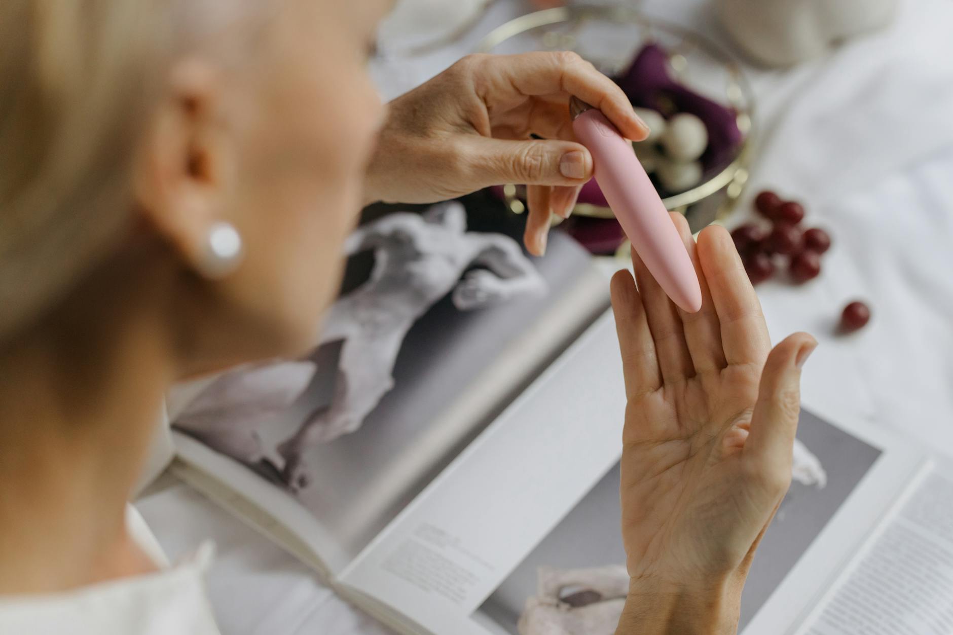 Woman holding a pink clitoral vibrator with art books in background, emphasizing intimate and intentional self-pleasure.