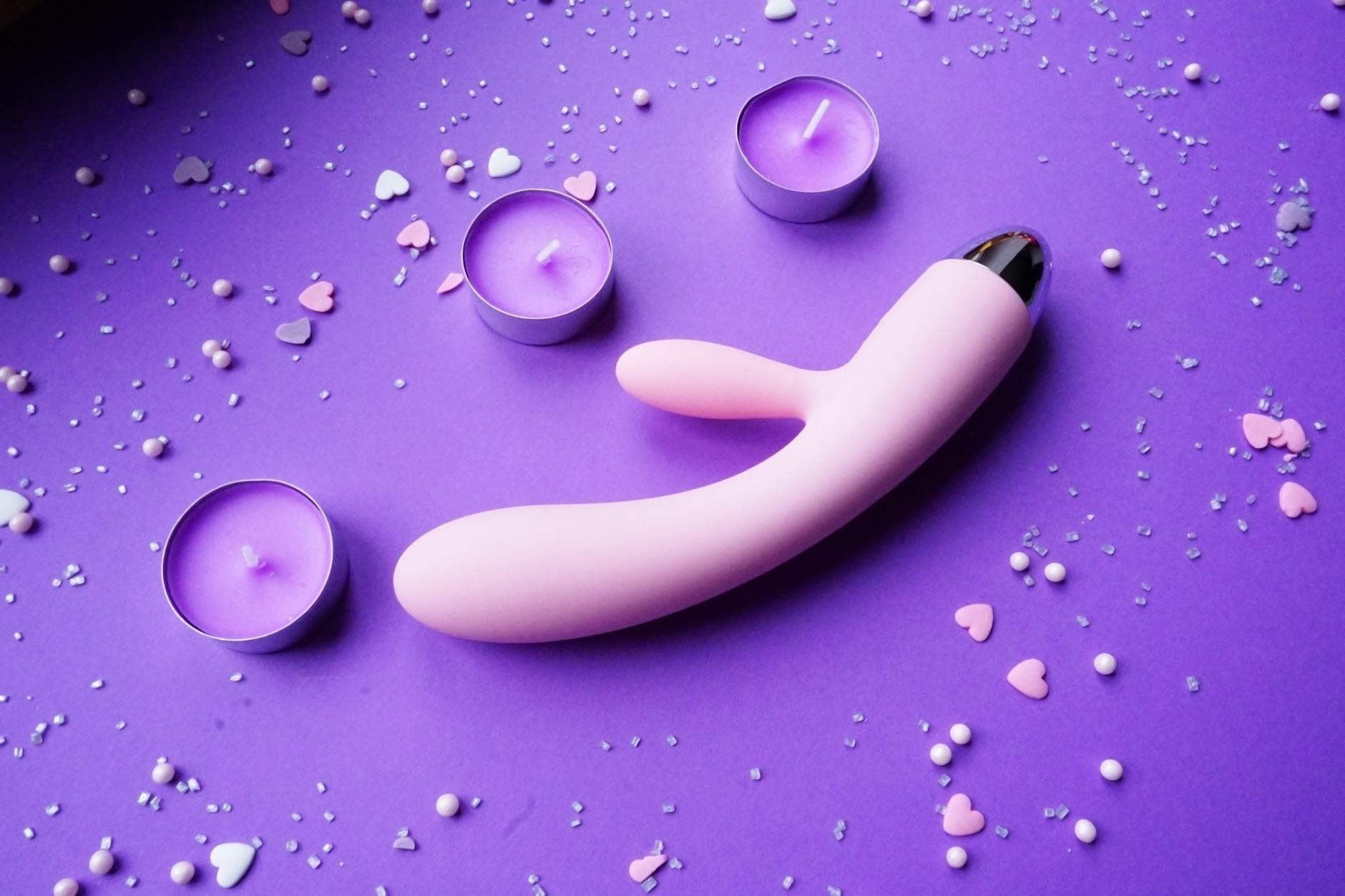 A pink clitoral vibrator on purple background with heart confetti and candles for a romantic couple setting